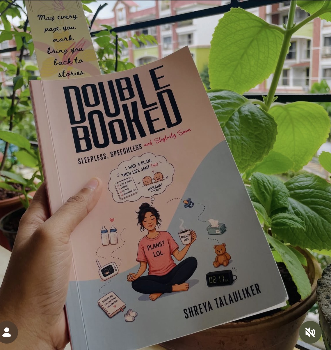 Reader holding Double Booked beside plants and a window